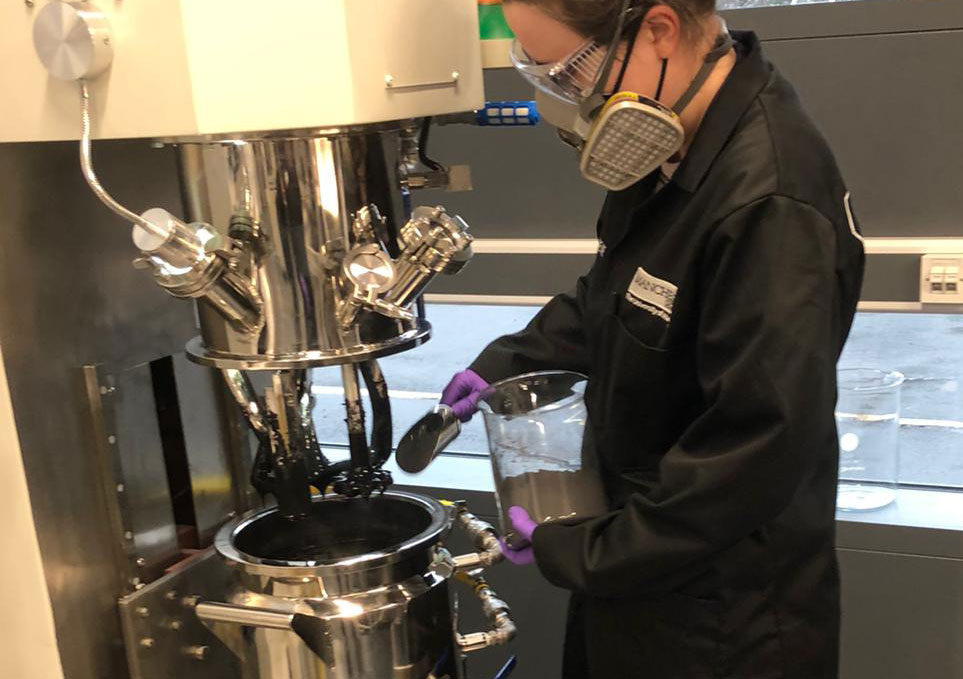 student in a lab coat scooping black granules into a large mixer
